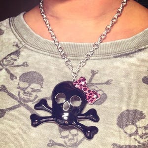 May include: A silver-toned chain necklace featuring a large, glossy black skull and crossbones pendant. The skull is adorned with a small, pink and black leopard-print bow detail attached to the upper right side of the skull's head.