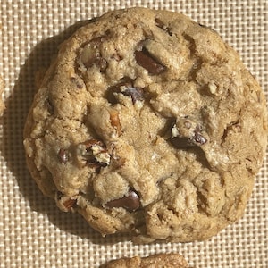 May include: Two golden brown chocolate chip cookies with pecans on a brown textured surface.