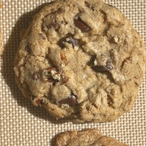 May include: Close-up of a large, golden brown chocolate chip cookie with pecans. The cookie is on a brown, textured baking sheet.