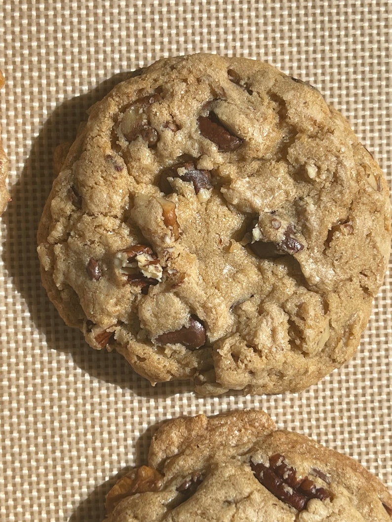 May include: Close-up of a large, golden brown chocolate chip cookie with pecans. The cookie is on a brown, textured baking sheet.