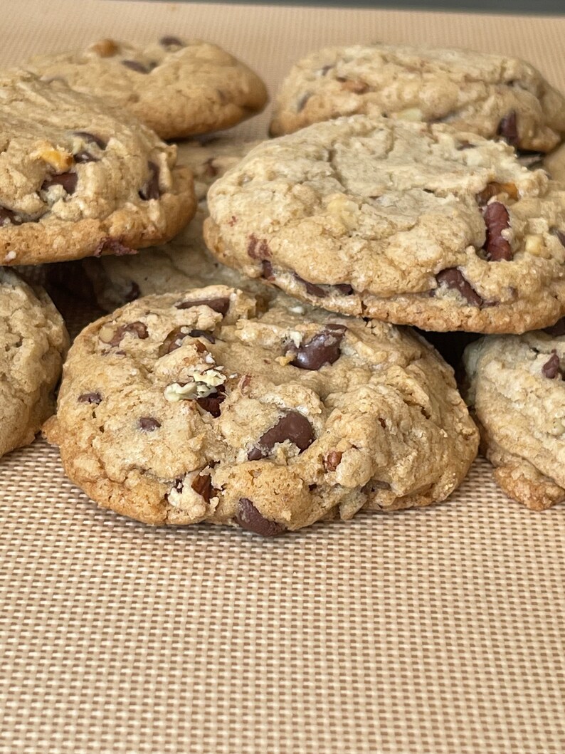 May include: A close-up of a stack of freshly baked chocolate chip cookies on a brown baking mat. The cookies are golden brown and have a slightly crispy texture.