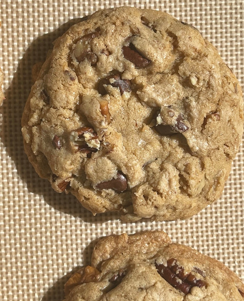 May include: Two golden brown chocolate chip cookies with pecans on a brown textured surface.