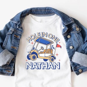 パーソナライズされたホールインワンの1歳の誕生日シャツ、カスタムゴルフの1歳の誕生日の家族お揃いのTシャツ、ゴルフカートの1歳の誕生日の男の子のパーティー衣装