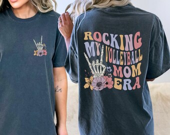 Może przedstawiać: Szary t-shirt z kwiatowym wzorem i napisem "Rocking My Volleyball Mom Era" z tyłu. Tekst jest w retro czcionce z kolorowymi literami.