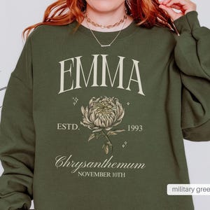 Peut inclure: Un sweat-shirt vert militaire avec le nom "EMMA" imprim&eacute; en blanc, un graphique de chrysanth&egrave;me et le texte "ESTD. 1993 Chrysanthemum NOVEMBER 10TH".