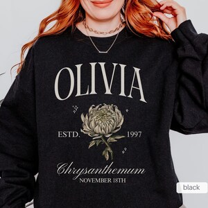 Peut inclure: Sweat-shirt noir avec le nom Olivia imprim&eacute; en blanc, un graphique de chrysanth&egrave;me stylis&eacute; et le texte "ESTD. 1997 Chrysanthemum NOVEMBER 18TH".