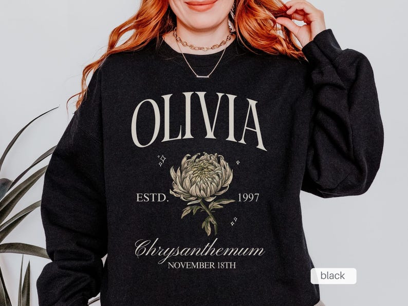 Peut inclure: Sweat-shirt noir avec le nom Olivia imprim&eacute; en blanc, un graphique de chrysanth&egrave;me stylis&eacute; et le texte "ESTD. 1997 Chrysanthemum NOVEMBER 18TH".