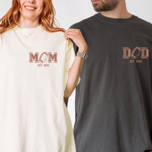 May include: Two t-shirts, one ivory and one pepper, with the words "MOM EST. 2025" and "DAD EST. 2025" printed on them. Each shirt features a football graphic.