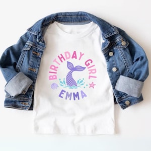 Pode incluir: Camiseta branca com um gráfico rosa, roxo e azul de uma cauda de sereia com o texto "Birthday Girl" e o nome "Emma" abaixo.
