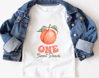 Peut inclure: T-shirt blanc avec un graphisme de pêche et le texte "ONE Sweet Peach" en corail et vert. Une veste en jean est drapée sur le t-shirt. L'illustration de la pêche est de style aquarelle avec une tige et des feuilles.