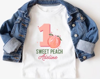 Peut inclure: T-shirt blanc avec un numéro un rose et une illustration de pêche. Le texte "Sweet Peach Adeline" est imprimé sous la pêche.