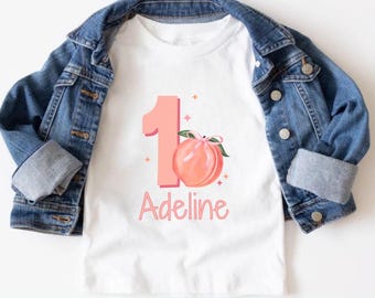Peut inclure: T-shirt à manches longues blanche avec un numéro un rose et un graphique de pêche. Le t-shirt porte le nom "Adeline" imprimé sous le numéro.