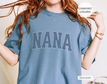 May include: A blue jean t-shirt with the word "NANA" printed in a large, white font. The shirt is a comfortable fit and has a relaxed, casual style.