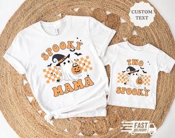 May include: Two white t-shirts with a Halloween theme. The larger shirt says "Spooky Mama" with a ghost wearing a witch hat and holding a pumpkin. The smaller shirt says "Two Spooky" with a ghost wearing a witch hat and holding a pumpkin.