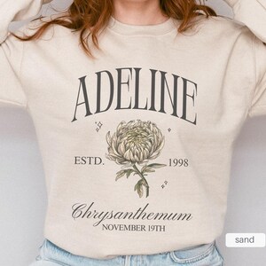 Peut inclure: Un sweat &agrave; manches longues de couleur cr&egrave;me avec le texte "Adeline Est. 1998" et un graphique d'une fleur de chrysanth&egrave;me avec le texte "Chrysanthemum November 19th".