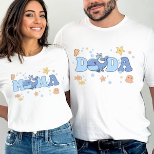 Op de afbeelding: Witte T-shirts met blauwe tekst en zeedierafbeeldingen. Eén shirt zegt "MAMA" met een walvis, de andere zegt "DADA" met een walvis. Beide shirts hebben zeesterren, vissen en bellen.