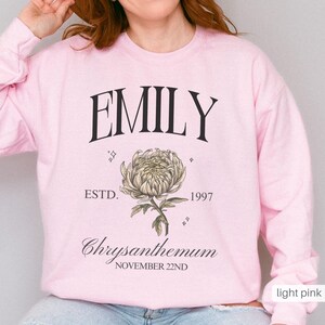 Peut inclure: Sweat-shirt rose p&acirc;le avec le nom "EMILY" imprim&eacute; en noir, ainsi que le texte "ESTD. 1997" et "Chrysanthemum NOVEMBER 22ND". Une illustration stylis&eacute;e d'une fleur de chrysanth&egrave;me est imprim&eacute;e sous le nom.