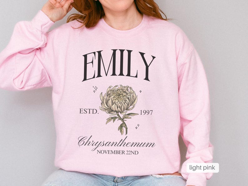 Peut inclure: Sweat-shirt rose p&acirc;le avec le nom "EMILY" imprim&eacute; en noir, ainsi que le texte "ESTD. 1997" et "Chrysanthemum NOVEMBER 22ND". Une illustration stylis&eacute;e d'une fleur de chrysanth&egrave;me est imprim&eacute;e sous le nom.
