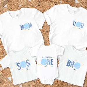 Puede incluir: Un conjunto de cinco camisetas blancas con diseños espaciales azules y amarillos. Las camisetas dicen "Mamá", "Papá", "Hermano", "Sos" y "Blasting into One".
