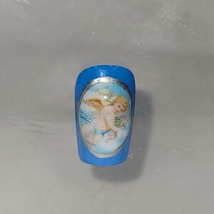 May include: A blue artificial nail with a silver oval frame containing a painted image of a cherub with wings, holding a bouquet of flowers.