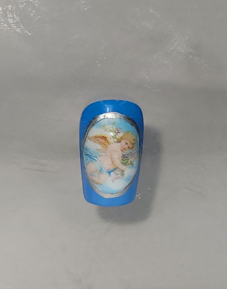 May include: A blue artificial nail with a silver oval frame containing a painted image of a cherub with wings, holding a bouquet of flowers.