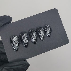 May include: Five black press-on nails with white abstract designs. The nails are arranged in a row on a black surface.