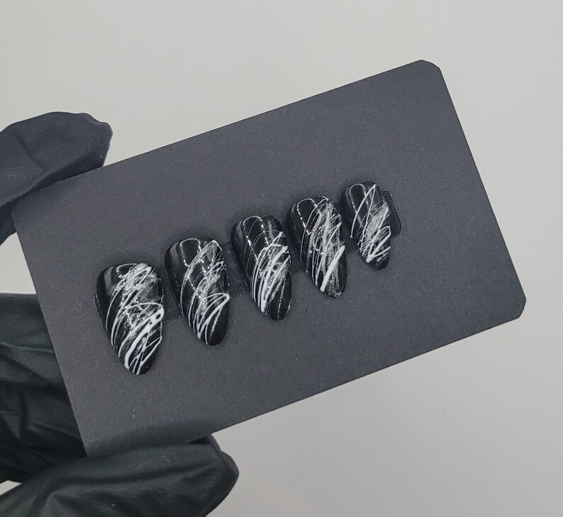 May include: Five black press-on nails with white abstract designs. The nails are arranged in a row on a black surface.