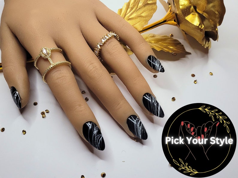 May include: Black and white swirl nail art on long, almond-shaped nails. The nails are painted black with white swirls and lines. The nails are on a hand wearing gold rings. The image also includes a black circle with white text that says "Pick Your Style".