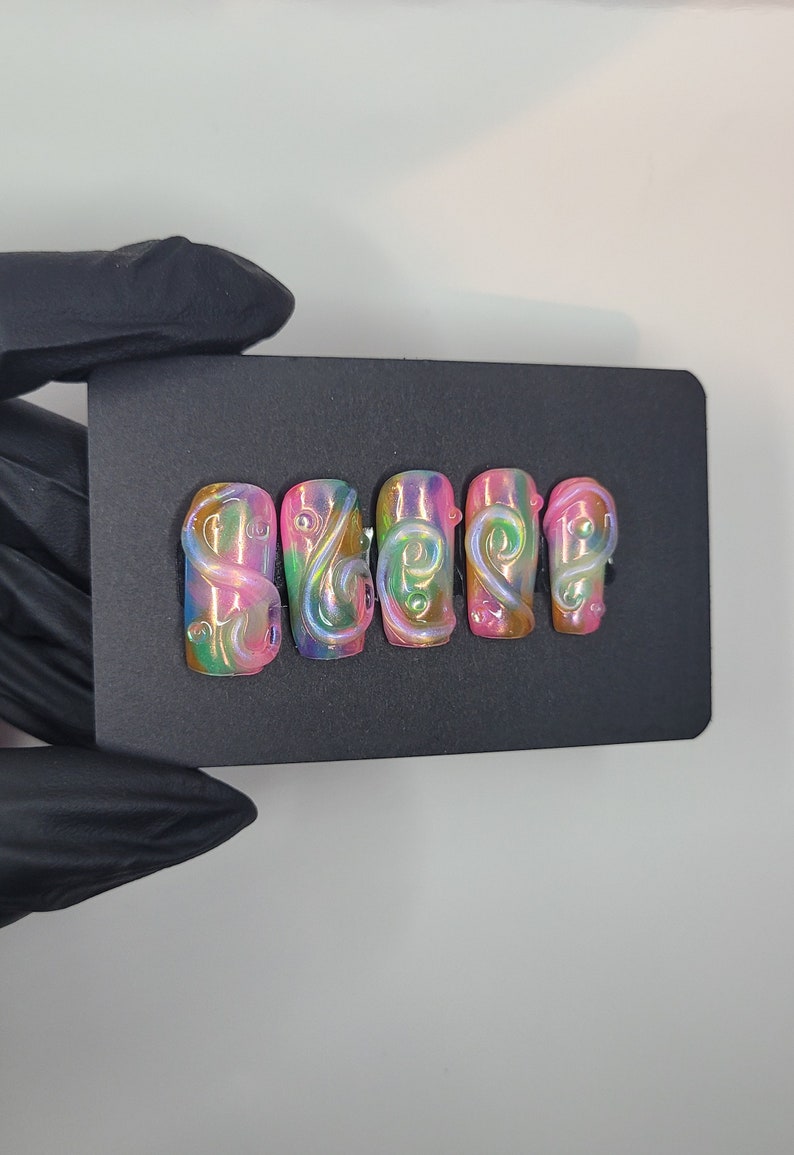 May include: Set of six false nails with a pink, green, and blue iridescent base and white swirl designs.