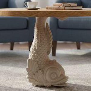 Op de afbeelding: Een kleine, ronde houten tafel met een visvormige voet. De voet is crèmekleurig en het tafelblad is lichtbruin. Een kop en schotel en een stapel boeken staan op de tafel. Twee blauwe fauteuils staan op de achtergrond.
