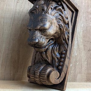 Puede incluir: Un ménsula de madera tallada con forma de cabeza de león en tonos marrones. La cara del león está detallada con una melena, nariz y pata. La ménsula tiene una parte superior rectangular y una base triangular.