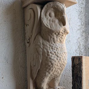 May include: A wooden owl sculpture with intricate details, carved in a realistic style. The owl is perched on a decorative base with scrollwork. The sculpture is made of light brown wood and has a natural finish.