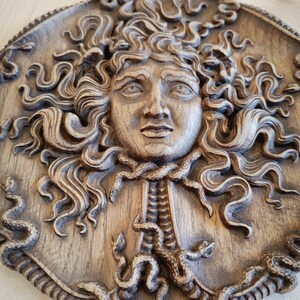 May include: A detailed, circular, wooden relief carving of Medusa's face, featuring intricately carved snakes for hair and a textured, aged appearance. The relief is in shades of brown and tan.