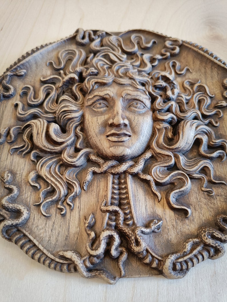 May include: A detailed, circular, wooden relief carving of Medusa's face, featuring intricately carved snakes for hair and a textured, aged appearance. The relief is in shades of brown and tan.