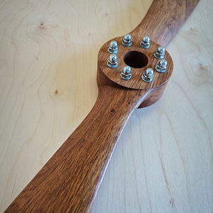 May include: A wooden airplane propeller with a brown stain finish. The propeller has a circular hub with eight silver screws around the center hole.