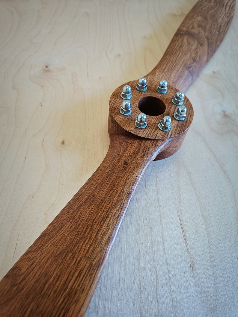 May include: A wooden airplane propeller with a brown stain finish. The propeller has a circular hub with eight silver screws around the center hole.