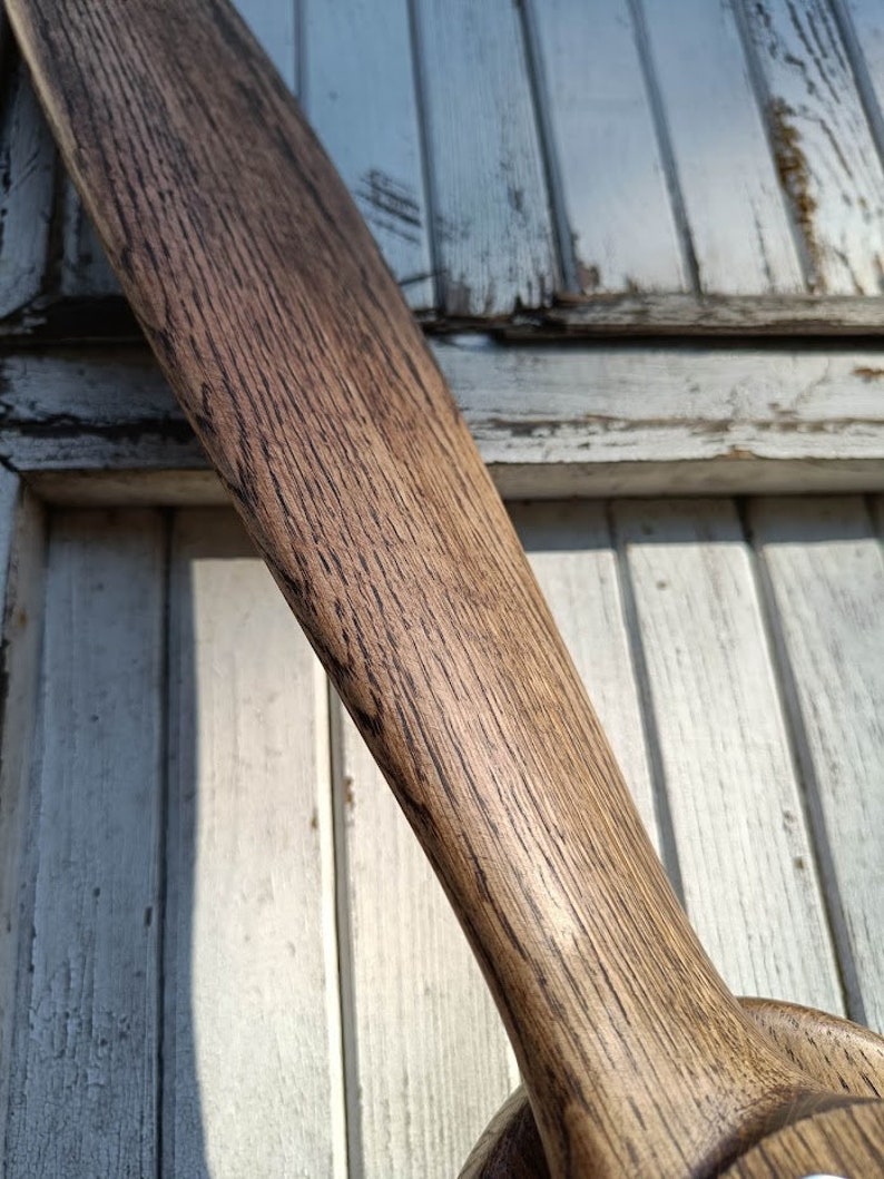 May include: A close-up of a wooden propeller with a dark brown stain. The propeller is made of wood with a grain pattern. The propeller is against a white wooden background.