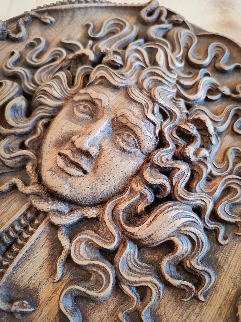 May include: A detailed, carved wooden relief depicting a classical face with flowing hair and snakes. The artwork is in shades of brown and gray, with intricate details in the hair and facial features. The relief is mounted on a wooden panel.
