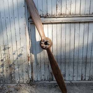 May include: A wooden airplane propeller with a brown finish and a silver metal hub. The propeller is leaning against a white wooden wall.