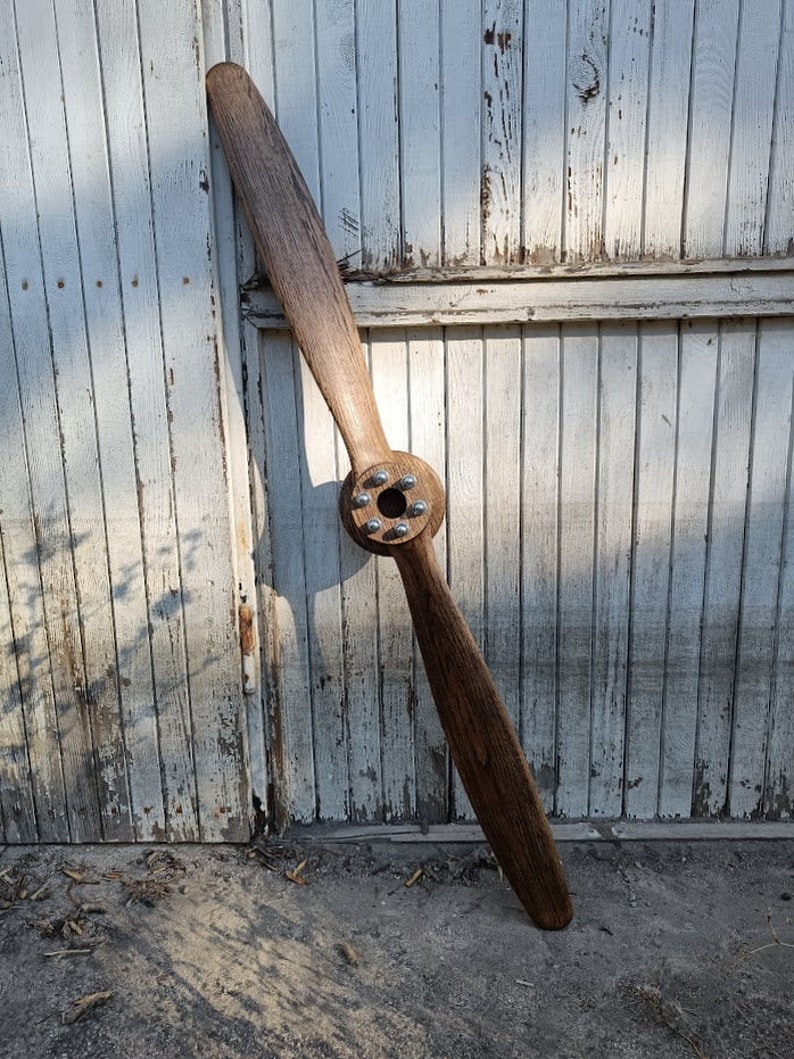 May include: A wooden airplane propeller with a brown finish and a silver metal hub. The propeller is leaning against a white wooden wall.