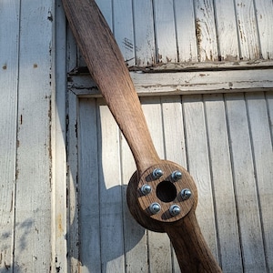 May include: A wooden airplane propeller with a brown finish and silver metal bolts. The propeller is mounted on a white wooden wall.