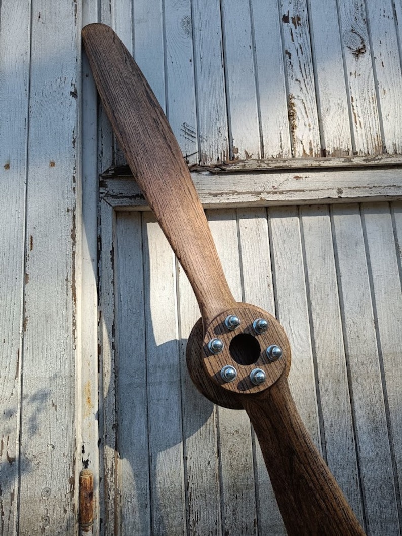 May include: A wooden airplane propeller with a brown finish and silver metal bolts. The propeller is mounted on a white wooden wall.