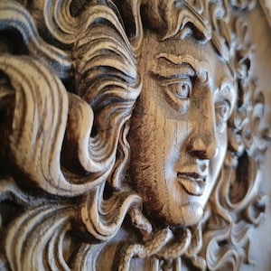 May include: A detailed, close-up of a carved wooden relief sculpture. The sculpture depicts a classical female face with flowing hair. The wood has a natural, aged appearance with visible grain and texture. The relief is in shades of brown and tan.