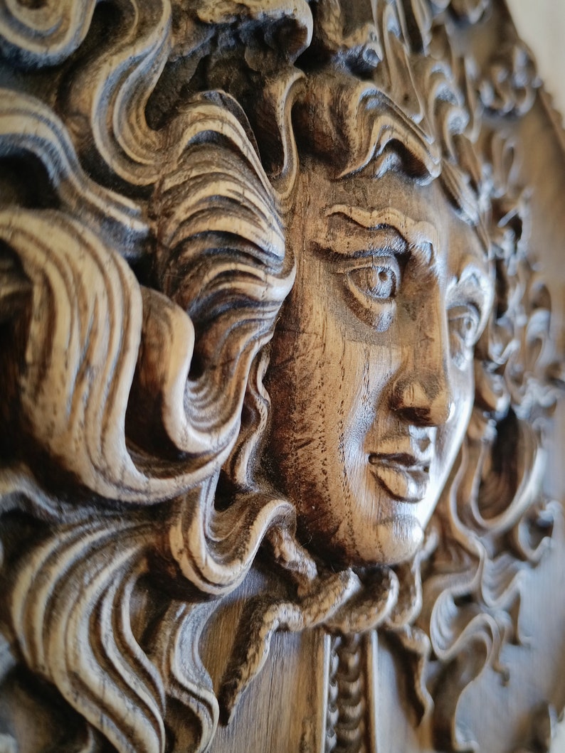 May include: A detailed, close-up of a carved wooden relief sculpture. The sculpture depicts a classical female face with flowing hair. The wood has a natural, aged appearance with visible grain and texture. The relief is in shades of brown and tan.