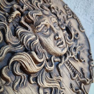 May include: A detailed, carved wooden relief depicting a classical female face with flowing hair and snakes. The relief is in shades of brown and tan, with intricate details in the hair and facial features. The artwork is mounted on a circular wooden plaque.