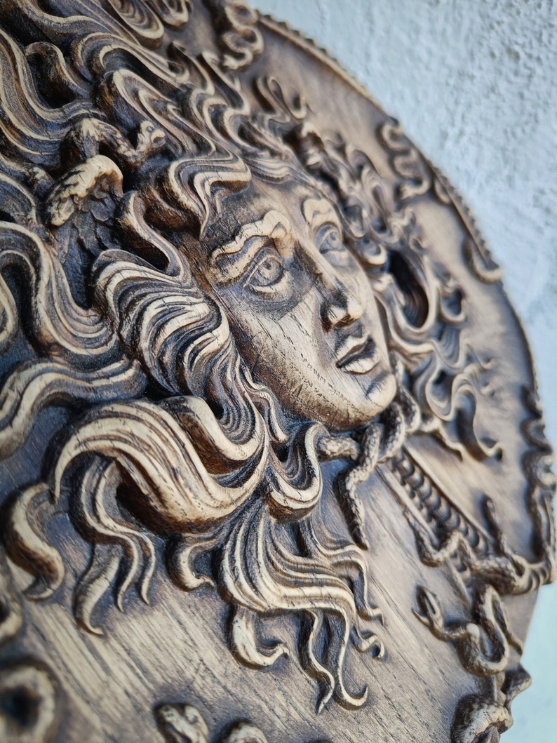 May include: A detailed, carved wooden relief depicting a classical female face with flowing hair and snakes. The relief is in shades of brown and tan, with intricate details in the hair and facial features. The artwork is mounted on a circular wooden plaque.