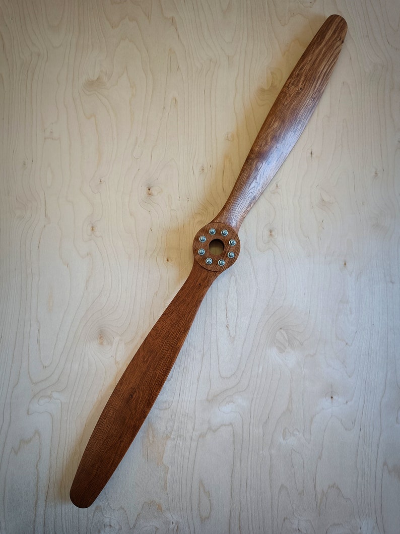 May include: A wooden airplane propeller with a brown finish and silver screws. The propeller is on a light wood background.