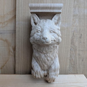 Puede incluir: Escultura de madera tallada con la cabeza de un zorro, con detalles de pelaje y rasgos faciales. El zorro es de color claro y parece ser un elemento arquitectónico decorativo, posiblemente un ménsula o soporte. La escultura está montada sobre una superficie de madera.