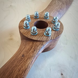 May include: A wooden propeller with a circular center and eight silver nuts around the edge. The propeller is brown and has a natural wood grain.