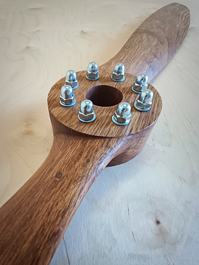 May include: A wooden propeller with a circular center and eight silver nuts around the edge. The propeller is brown and has a natural wood grain.